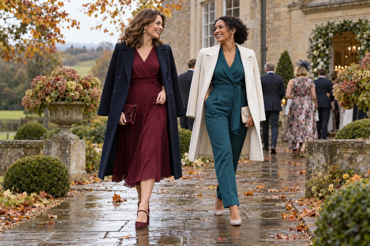 Two women in a burgundy dress and teal jumpsuit with coats at a UK autumn wedding